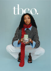 Woman wearing a long skinny merino wool red scarf with green jelly on a silver dish, with a light blue background, featuring the brand 'tbco' logo.