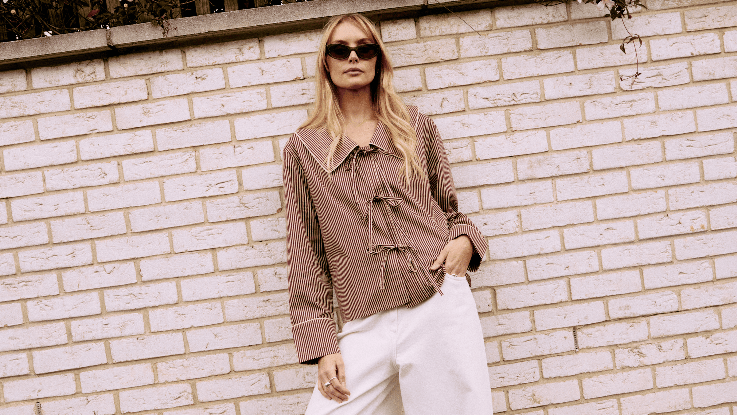 Woman wearing a TBCo camel stripe shirt and white pants against a light brick wall.