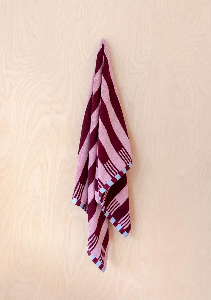 TBCo Terry Cotton Towels in Burgundy Stripe hanging flat against pale wood wall showing bold pink and burgundy pattern