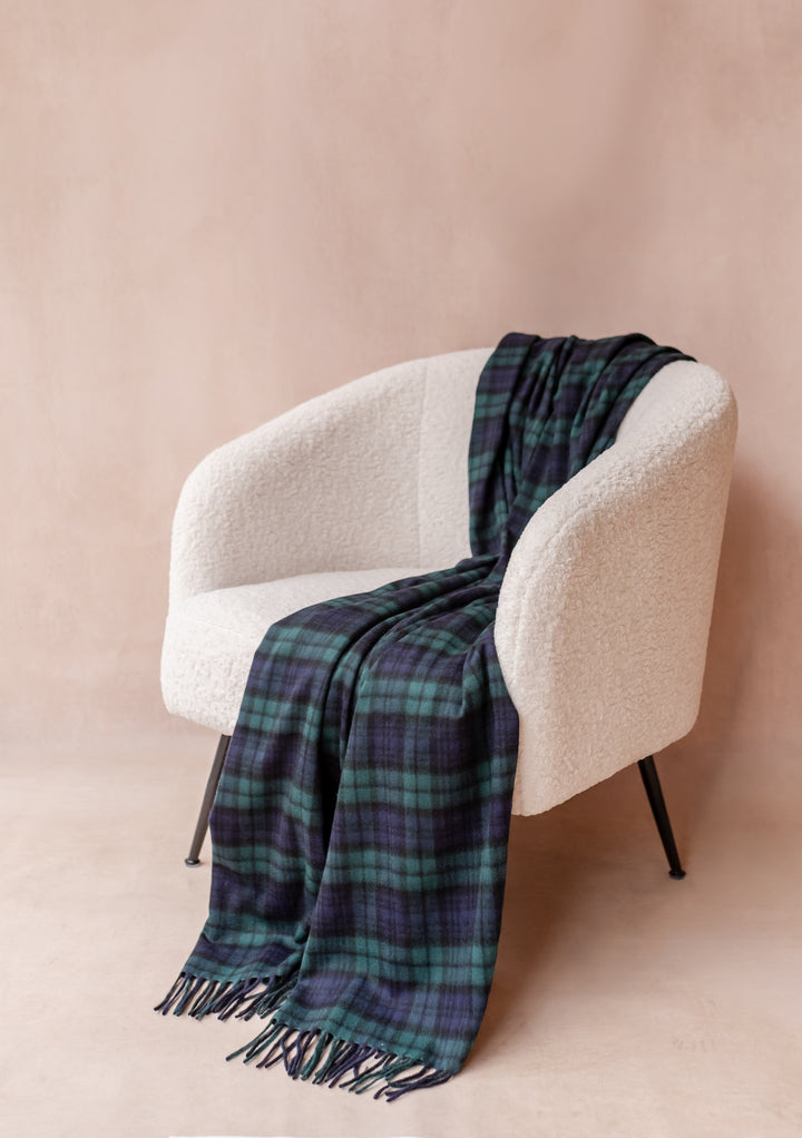 Cashmere Blanket in Black Watch Tartan by TBCo draped over a cream boucle armchair with fringe detailing.