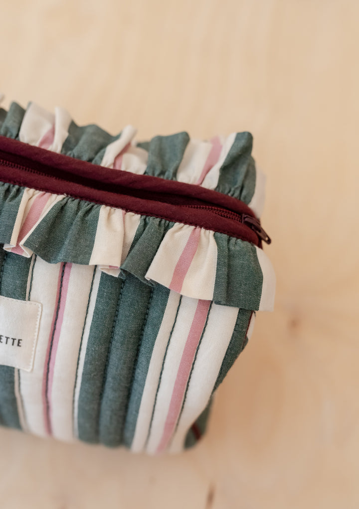 TBCo x Glassette Cotton Wash Bag in Green Stripe, featuring quilted fabric, burgundy zipper, ruffled trim, and multiple interior compartments on a light wood surface.