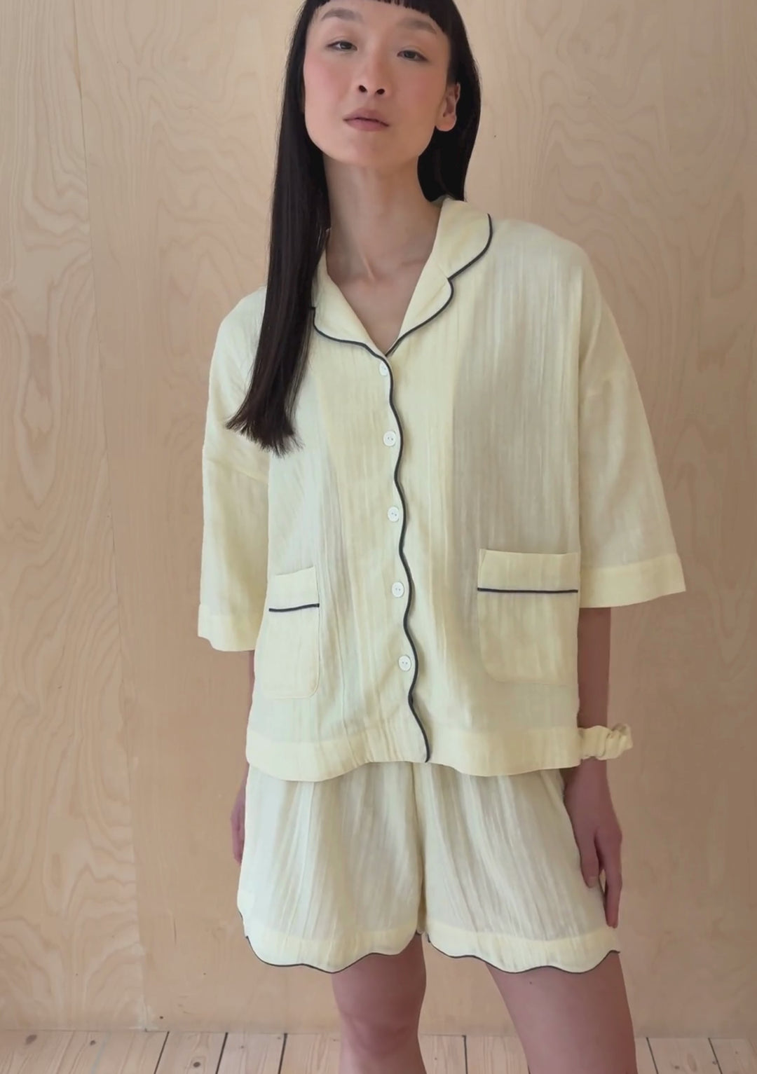A model poses against a neutral ply backdrop, wearing a buttermilk yellow TBCo pyjama shorts and shirt set.